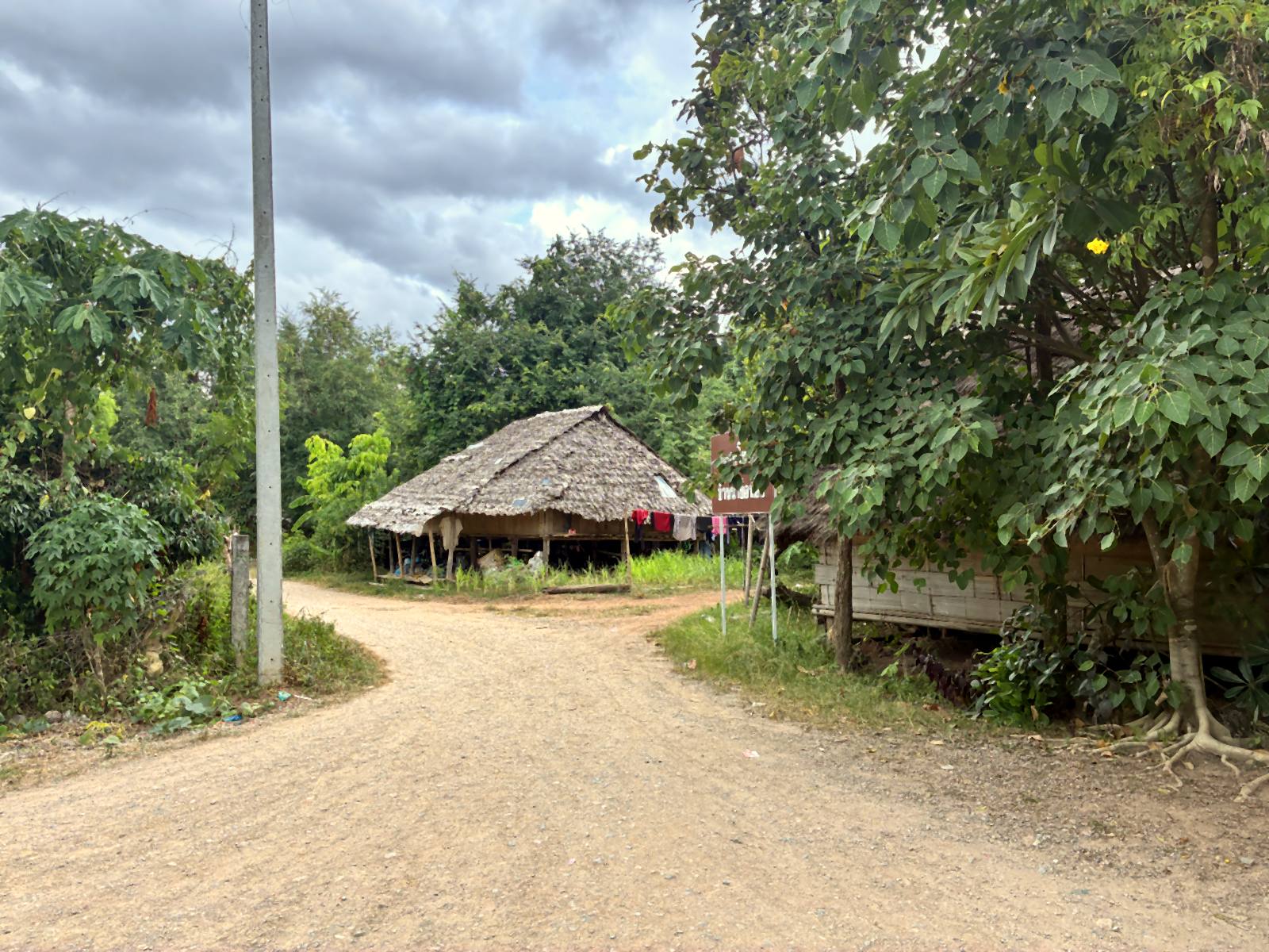 A concrete power pole at the roadside of a Karen village in Lamphun, with the thatched roofs of Ban Nam Bo Noi just beyond it